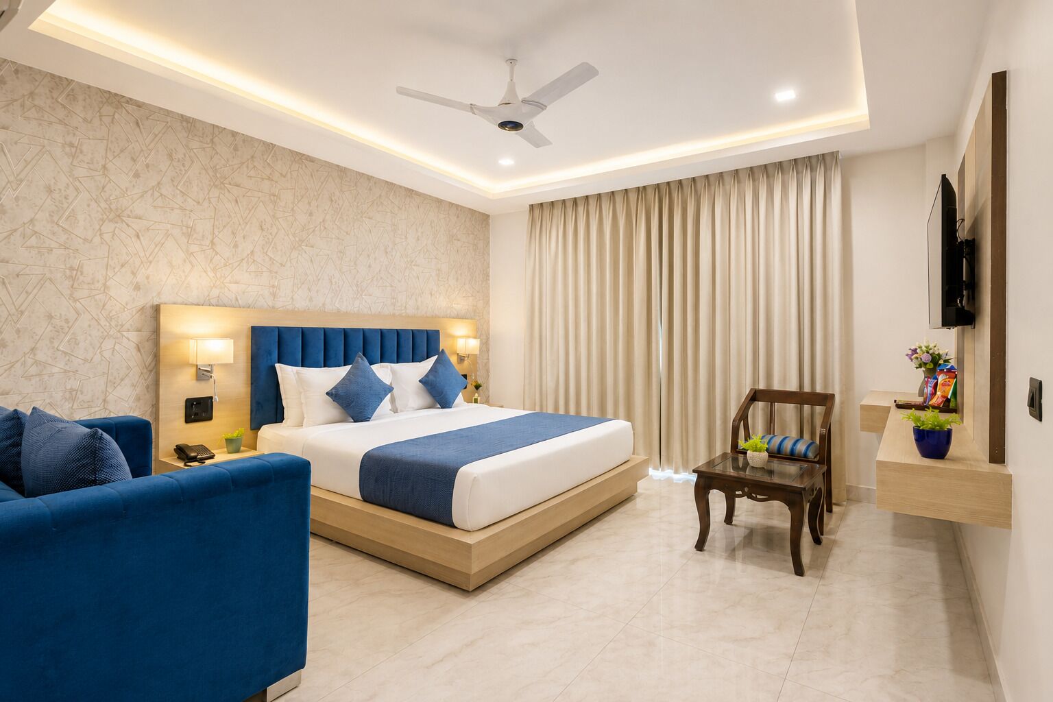 A hotel room with a bed, a blue sofa, a wooden nightstand, a desk, a chair, a ceiling fan, and a TV mounted on the wall.