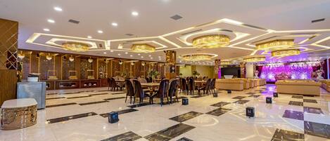 A spacious hotel lobby with a checkered floor, multiple dining tables, and elegant chandeliers.