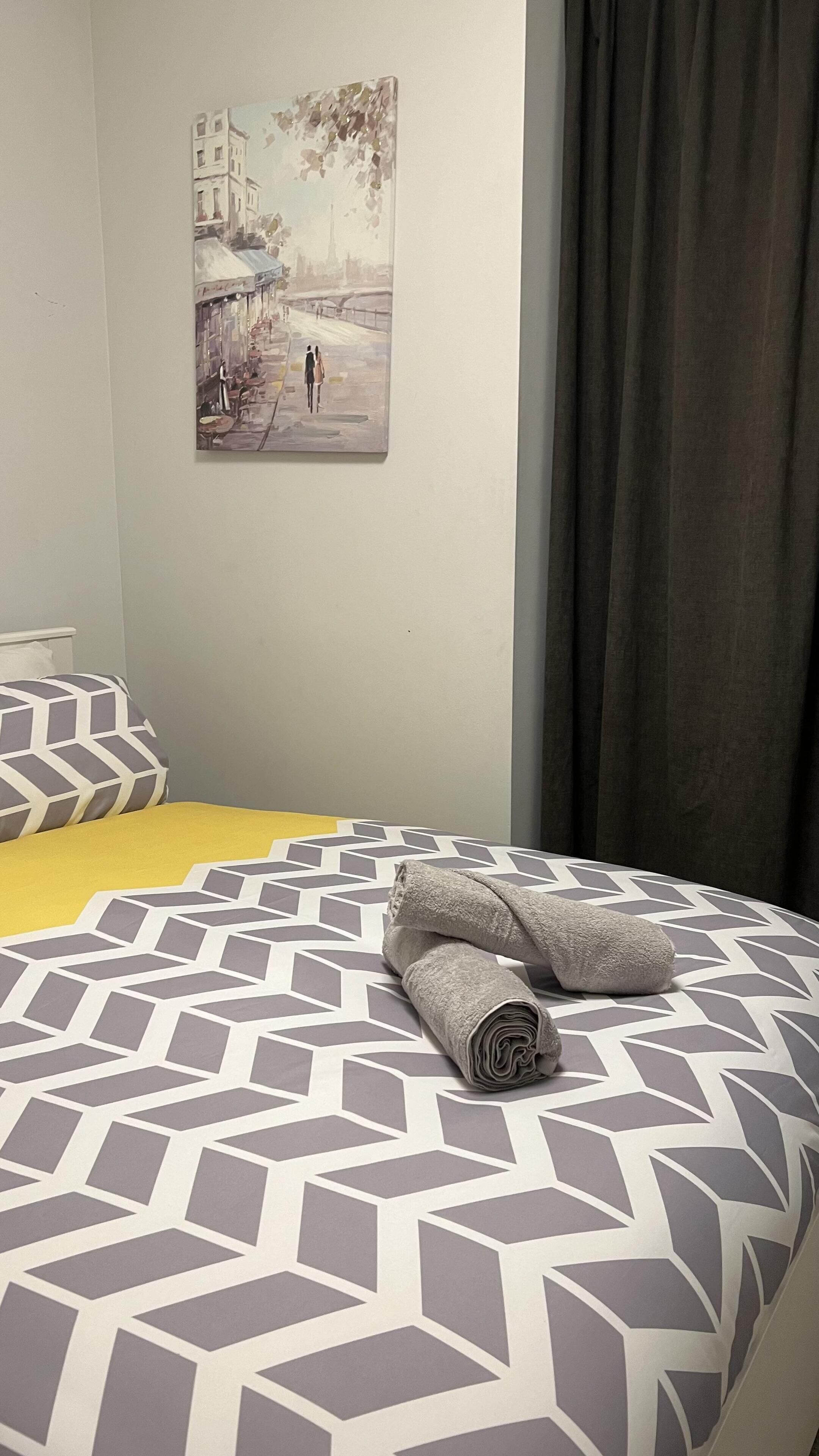 Una cama con una colcha de estampado geométrico, una toalla enrollada y un cuadro de un paisaje urbano en la pared.