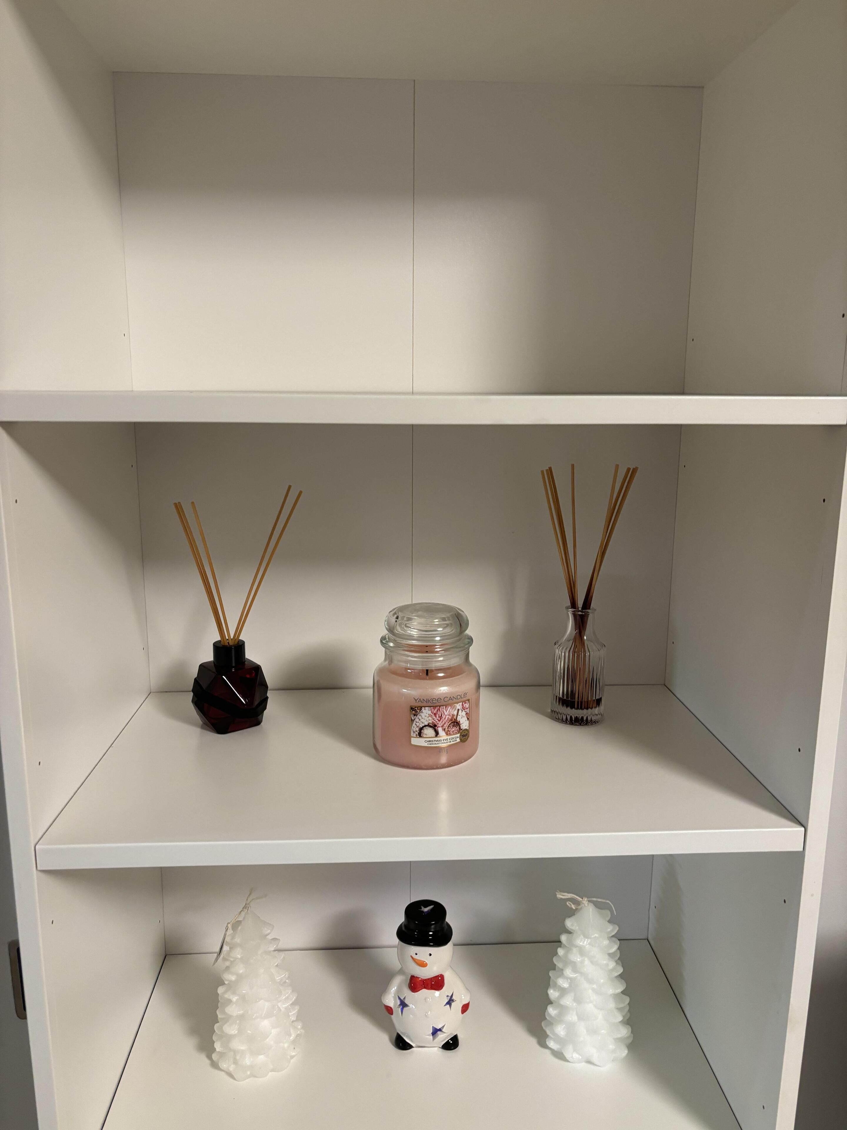 A white shelf with a pink candle, a black vase, and two glass vases with reed sticks.