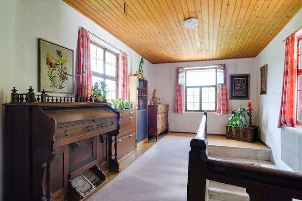 Une pièce avec un plafond en bois, un piano, une fenêtre ornée de rideaux rouges et un tableau accroché au mur.
