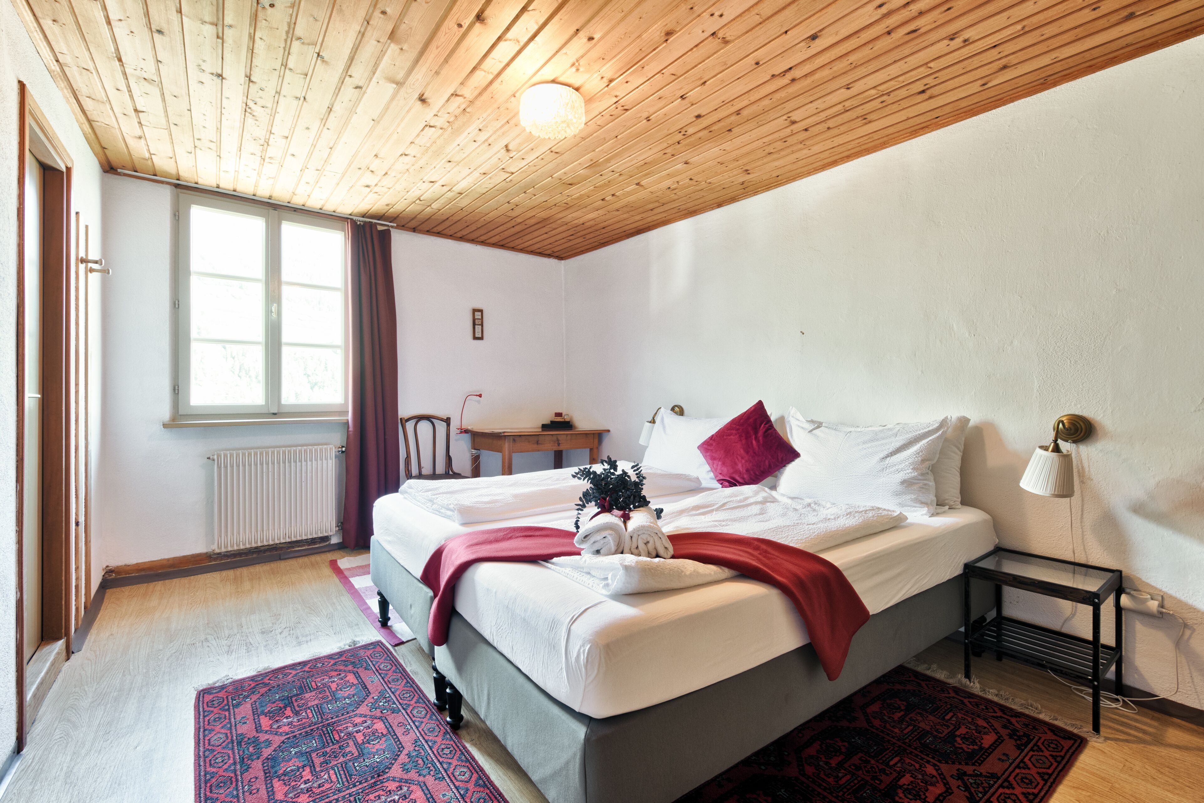 A bedroom with a wooden ceiling, a bed with white bedding and red accents, a small table, and a window with curtains.