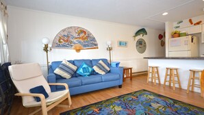 Sala de estar con un sofá azul, un sillón blanco, taburetes de madera, un tapiz de colores y una zona de cocina con un refrigerador y una calcomanía de pez.