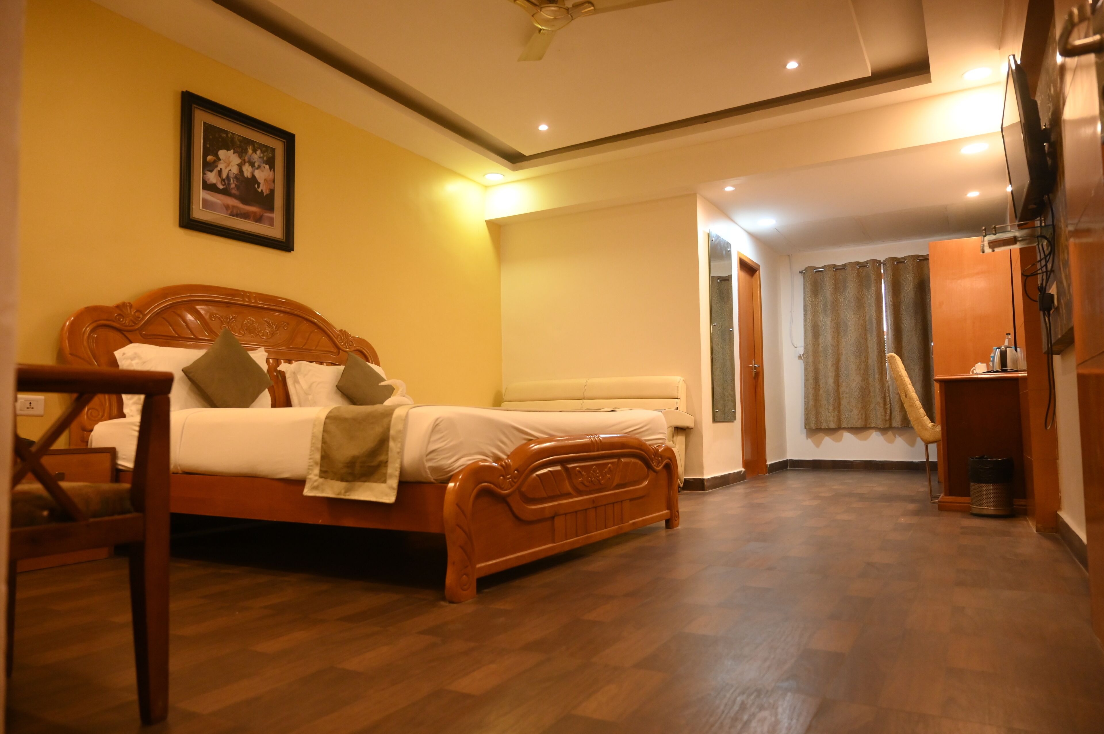Habitación de hotel con cama de madera, una silla, una mesita de noche, televisor y cortina.
