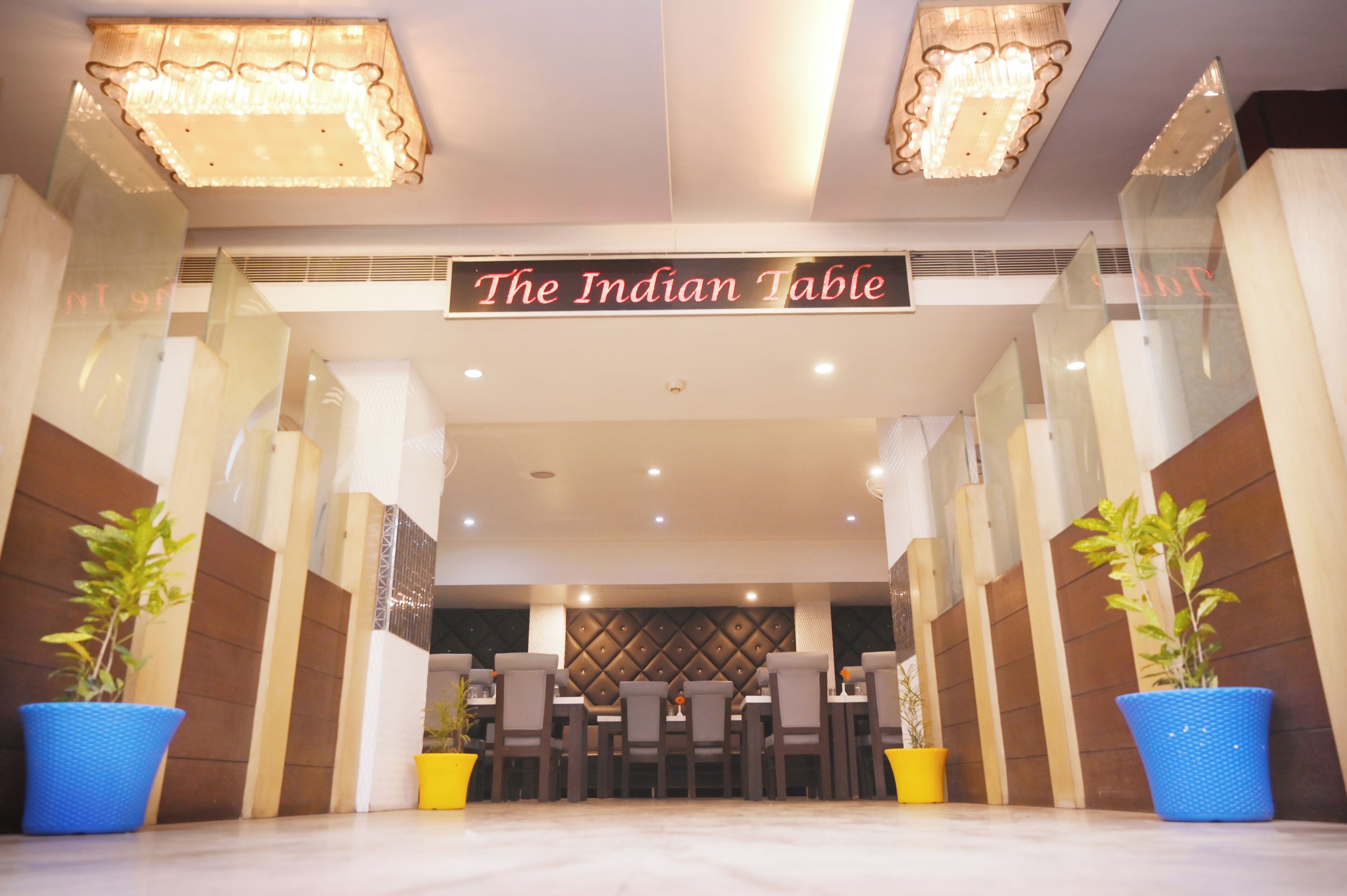 A restaurant entrance with a sign reading "The Indian Table," featuring a modern interior with a central dining area, potted plants, and a decorative ceiling.