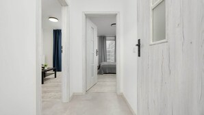 Un couloir doté d’un plancher de couleur claire, de murs blancs et d’une porte donnant sur une chambre à coucher avec un lit et des rideaux.