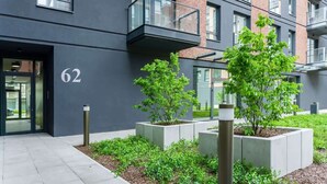 A modern building with a flat facade, numbered 62, featuring a balcony and a small garden area with shrubs and young trees.
