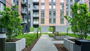A modern apartment building with a central courtyard, featuring a paved walkway, planters with greenery, and balconies.