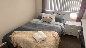 Una cama bien tendida con una manta gris y beige, una toalla blanca enrollada y una almohada estampada. Hay una mesita de noche con una lámpara y un cajón. La habitación tiene una ventana con persianas, una alfombra oscura y una pared lisa.