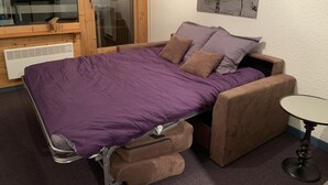 A compact bed with a purple mattress and brown headboard, a small bedside table, and a window with a view of trees.