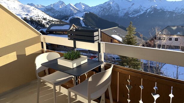 A balcony with a table and chairs, overlooking snow-covered mountains and a village.