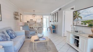 A spacious living room with a sofa, coffee table, and a TV on a stand. There is a dining area with chairs and a table in the background.