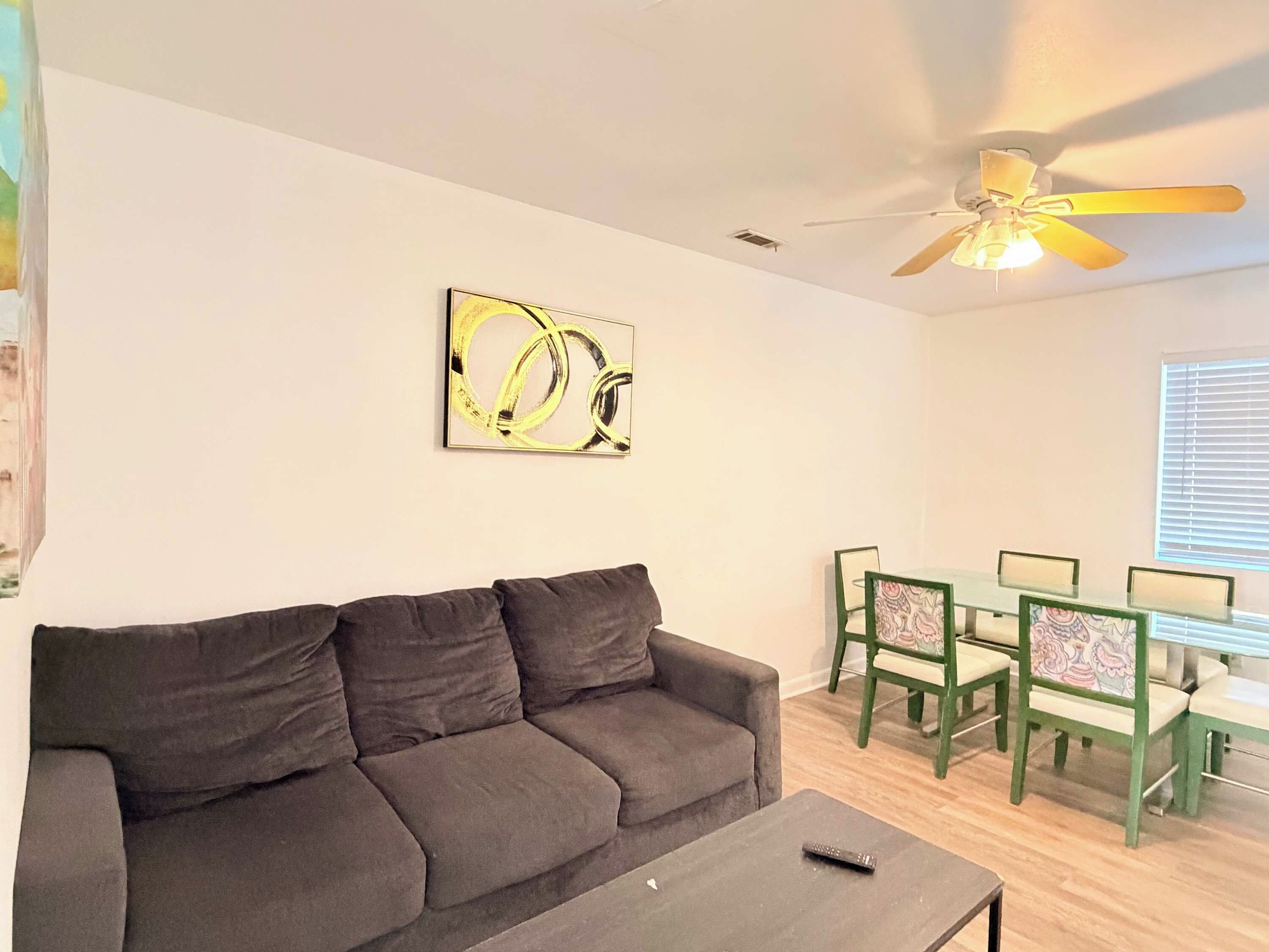 A living room with a sofa, a coffee table, a ceiling fan, a dining table with chairs, and a wall-mounted artwork.
