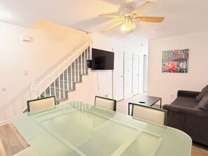 A modern living room with a glass-top dining table, a gray sofa, a ceiling fan, and a staircase leading to another room. - Beautiful and Tranquil Cherry Grove Beach (North Myrtle Beach)