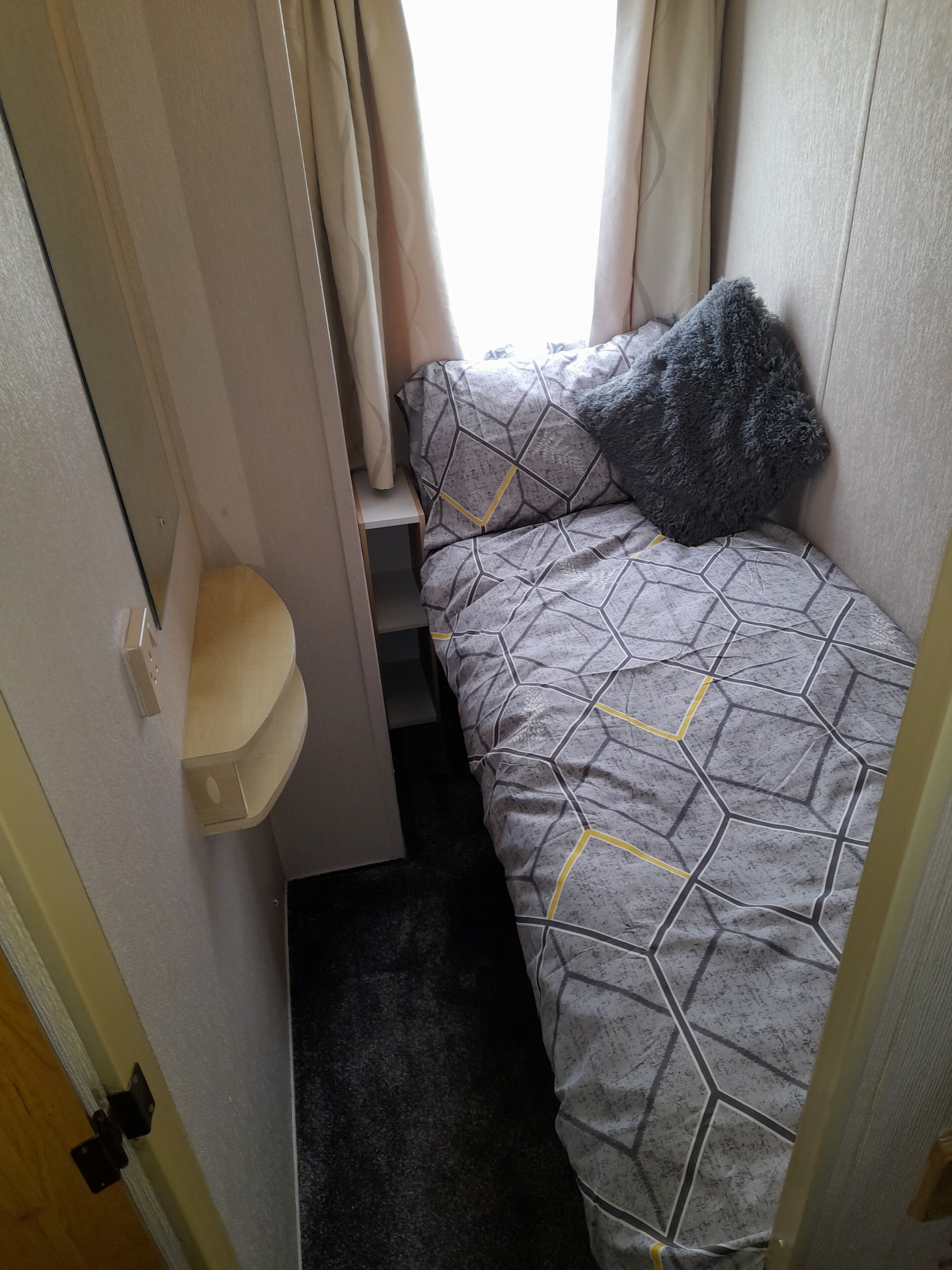 A small, narrow room with a bed, a small shelf, and a window with curtains.