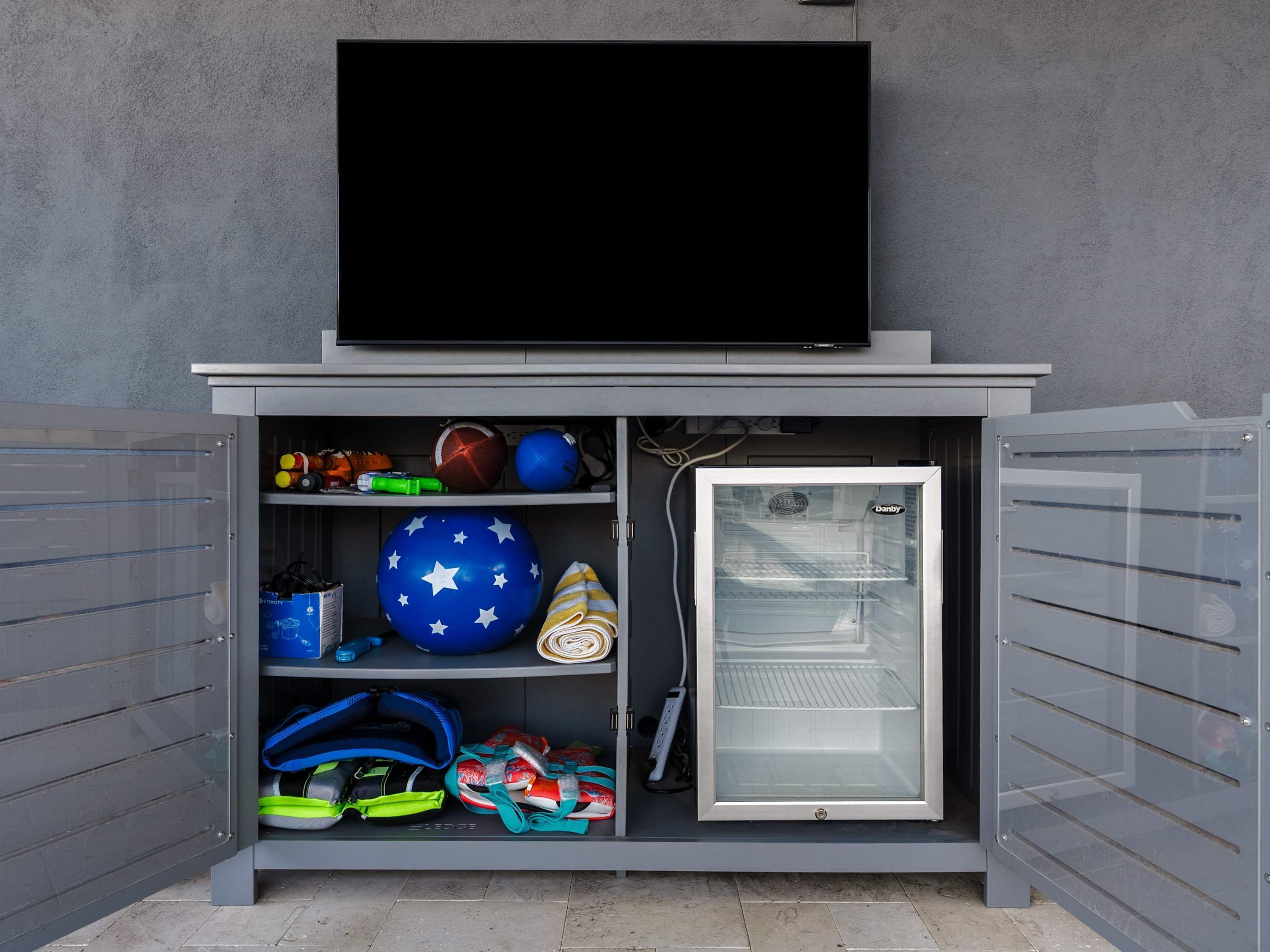 A compact storage unit with a television mounted above, containing sports balls, a cooler, and various bags.