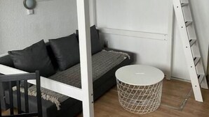 Ein kleines Zimmer mit Etagenbett, einem kleinen Tisch, einem runden weißen Korb und einer an der Wand befestigten Leiter.