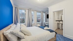 Een slaapkamer met een groot bed, meerdere ramen, een badkamer met een wastafel en spiegel, en een blauwe accentmuur.