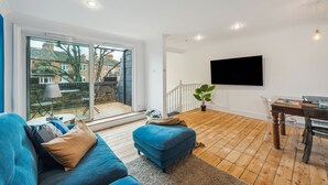 Een moderne woonkamer met een blauwe bank, een houten eettafel en een aan de muur bevestigde televisie.