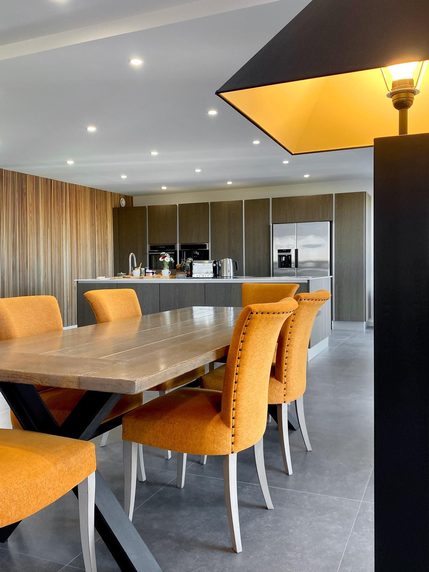 Een moderne keuken met een centraal eiland, roestvrijstalen apparatuur en een eetgedeelte met een houten tafel en oranje stoelen.