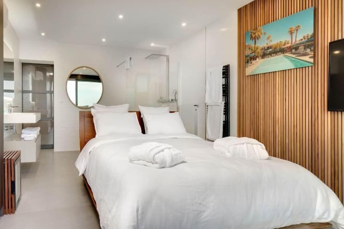 A modern hotel room with a large bed, a round mirror, a painting, and a TV on the wall.