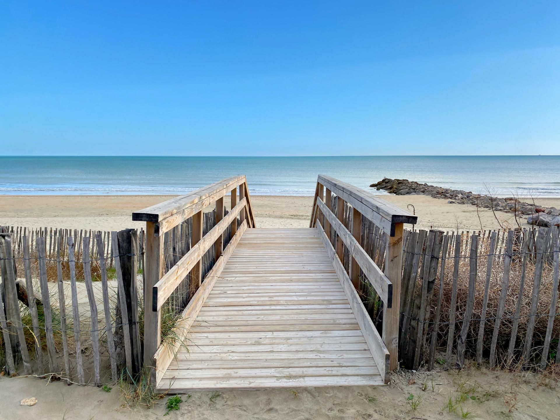 Een houten looppad dat naar een zandstrand leidt, onder een heldere blauwe lucht.