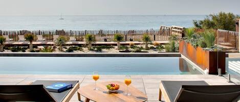 Zona de piscina con sillones reclinables, una mesa con frutas y bebidas, y vista al mar.