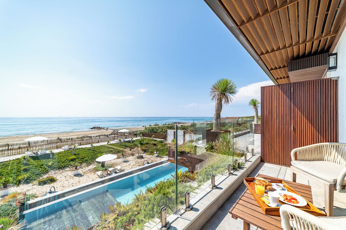 A poolside area with a view of the beach and ocean, a wooden deck, and a table with breakfast items.