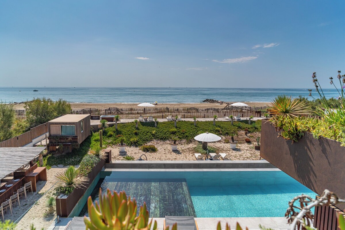 A poolside view with a beach and ocean in the background, wooden deck, and lounge chairs.
