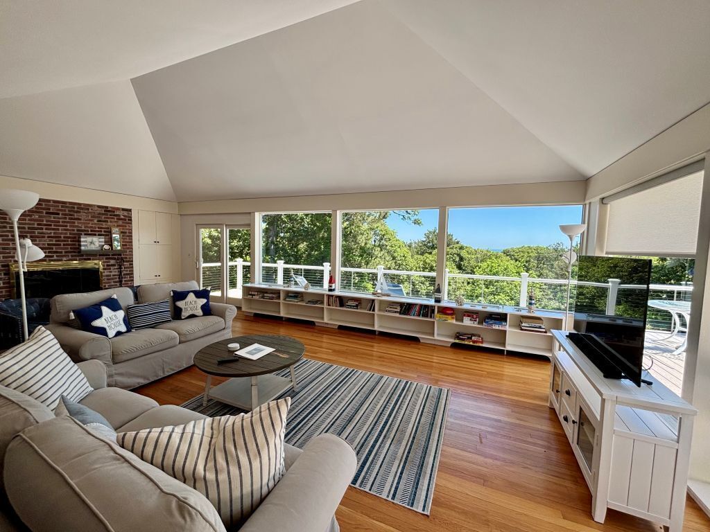 A spacious living room with a sectional sofa, a coffee table, and a fireplace. There is a balcony with a railing and a TV on a stand.