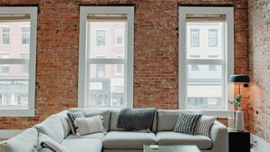 A living room with a brick wall, large windows, a sectional sofa, a coffee table, and a lamp.