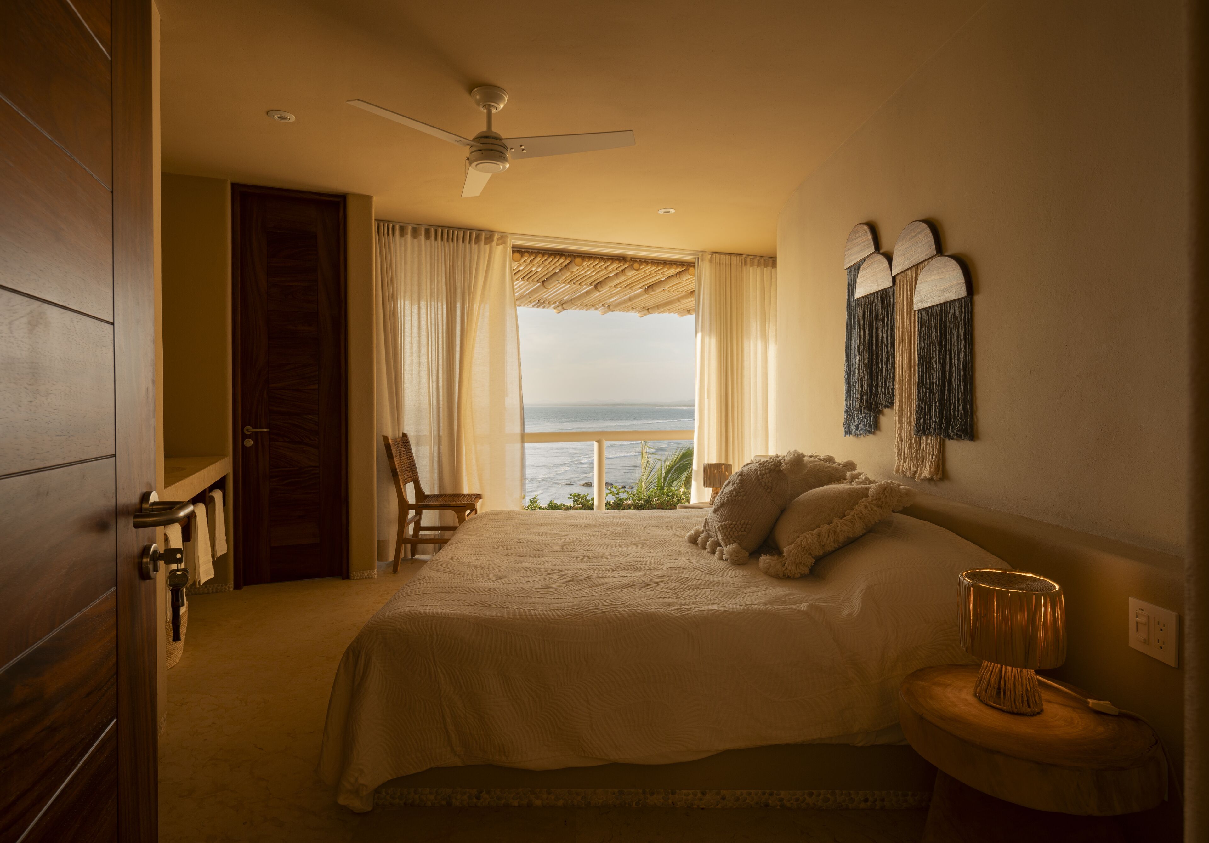 A bedroom with a large bed, a ceiling fan, a view of the ocean, and a small table with a lamp.