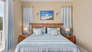 Une chambre à coucher avec un lit, deux tables de chevet, une tête de lit en bois, un tableau sur le thème de la plage et une fenêtre avec des rideaux.