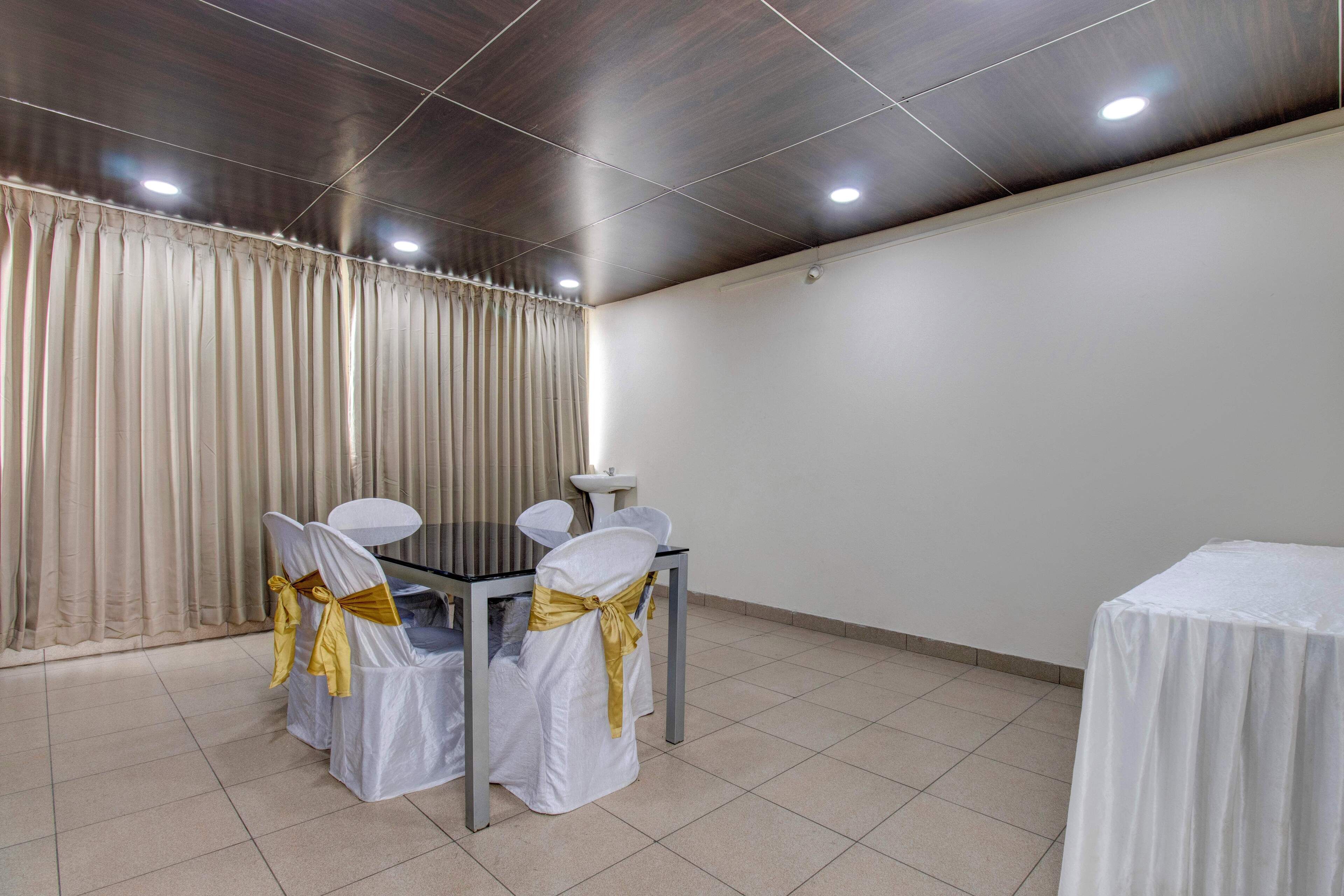 Un salón de conferencias con una mesa redonda, cuatro sillas con fundas blancas y cintas doradas, un mantel blanco, una cortina blanca y un piso de baldosas.