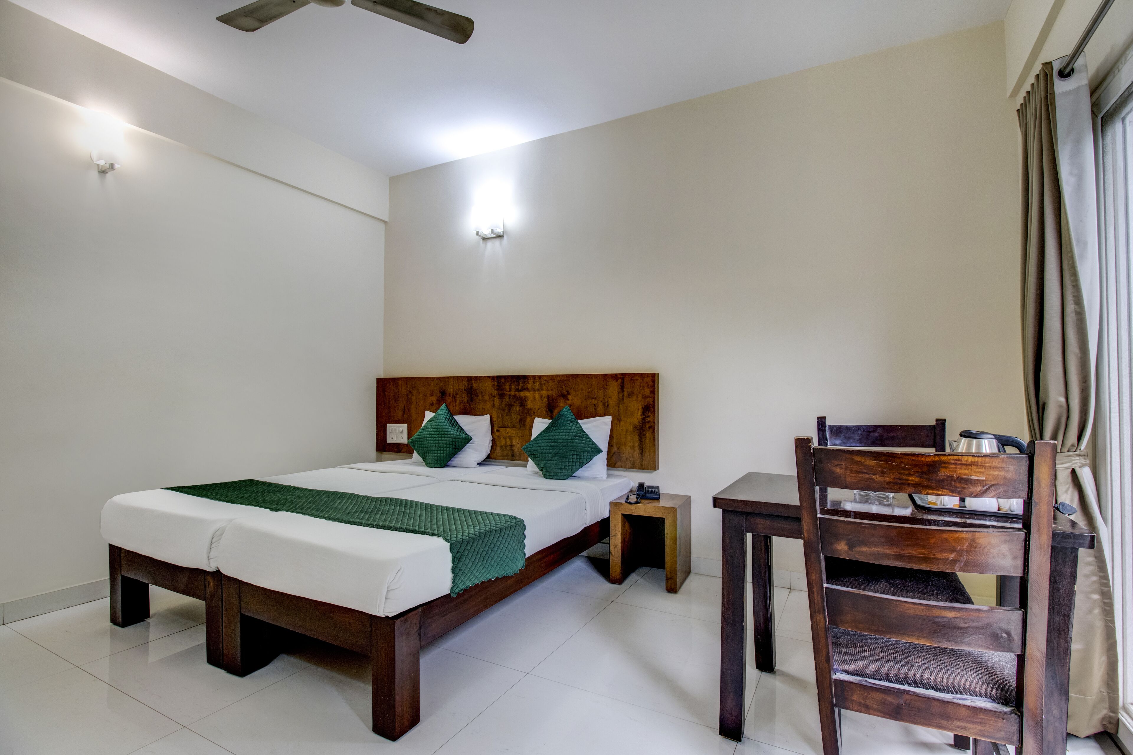 Una habitación de hotel ordenada con cama, mesita de noche, silla y una pequeña mesa con una tetera.