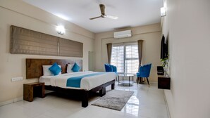 Une chambre à coucher comprenant un lit, des tables de chevet, un ventilateur de plafond, un appareil de climatisation, une fenêtre avec des rideaux et une petite table avec une chaise.