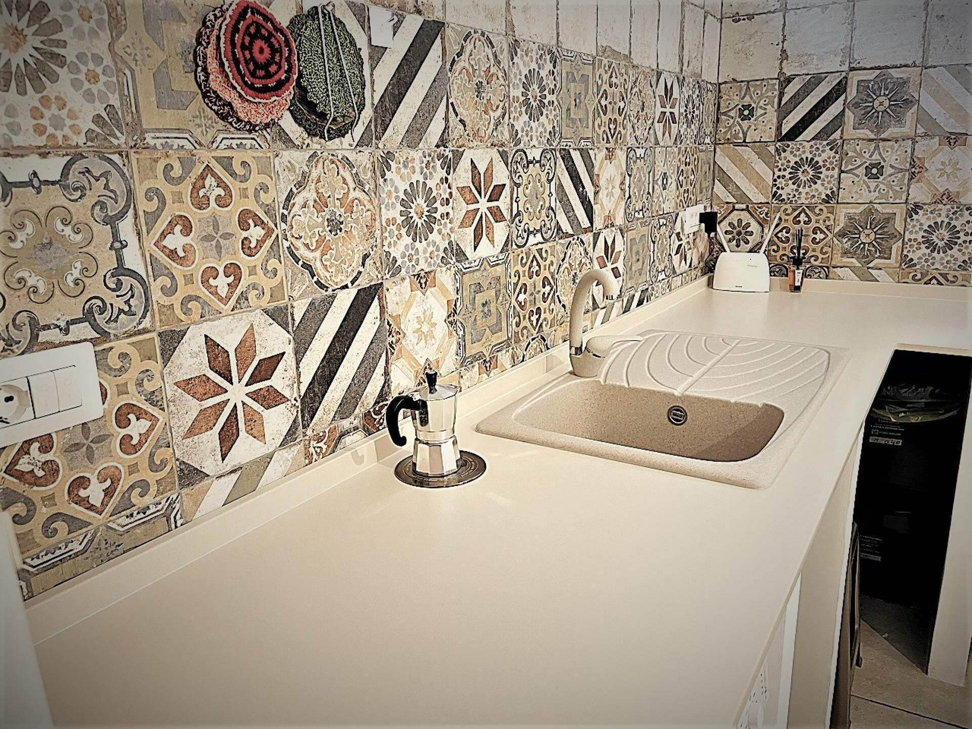 Una cocina con encimera blanca, cafetera y lavamanos. El salpicadero está hecho con azulejos de diversos diseños.