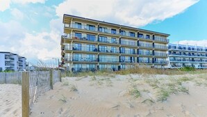 Un hotel de varios pisos con balcones, situado en una playa de arena con una cerca de madera al frente.