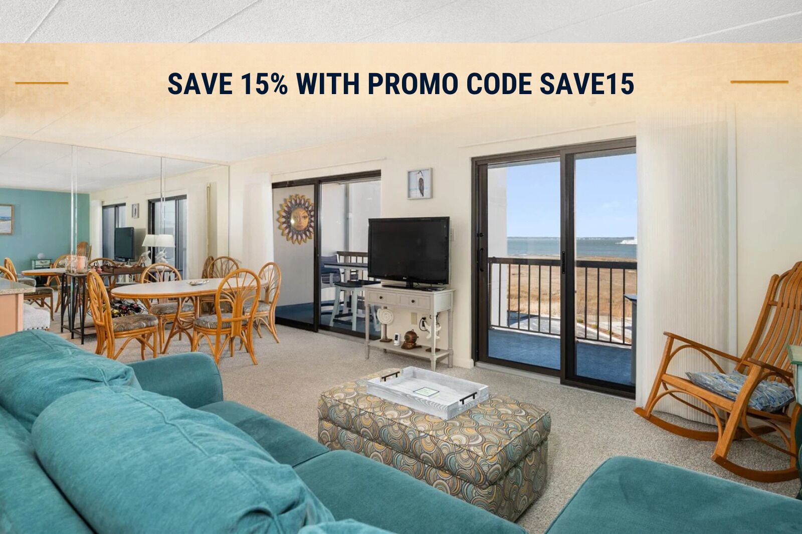 A living room with a sofa, ottoman, dining table, chairs, a television, and a balcony with a view of the beach.