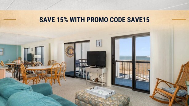 A living room with a sofa, ottoman, dining table, chairs, a television, and a balcony with a view of the beach.