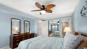 A bedroom with a bed, dresser, two mirrors, a ceiling fan, and a door leading to another room.