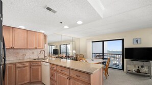 A modern kitchen with wooden cabinets, a granite countertop, and a large island. There is a dining area with a table and chairs, and a balcony with a view of the ocean.
