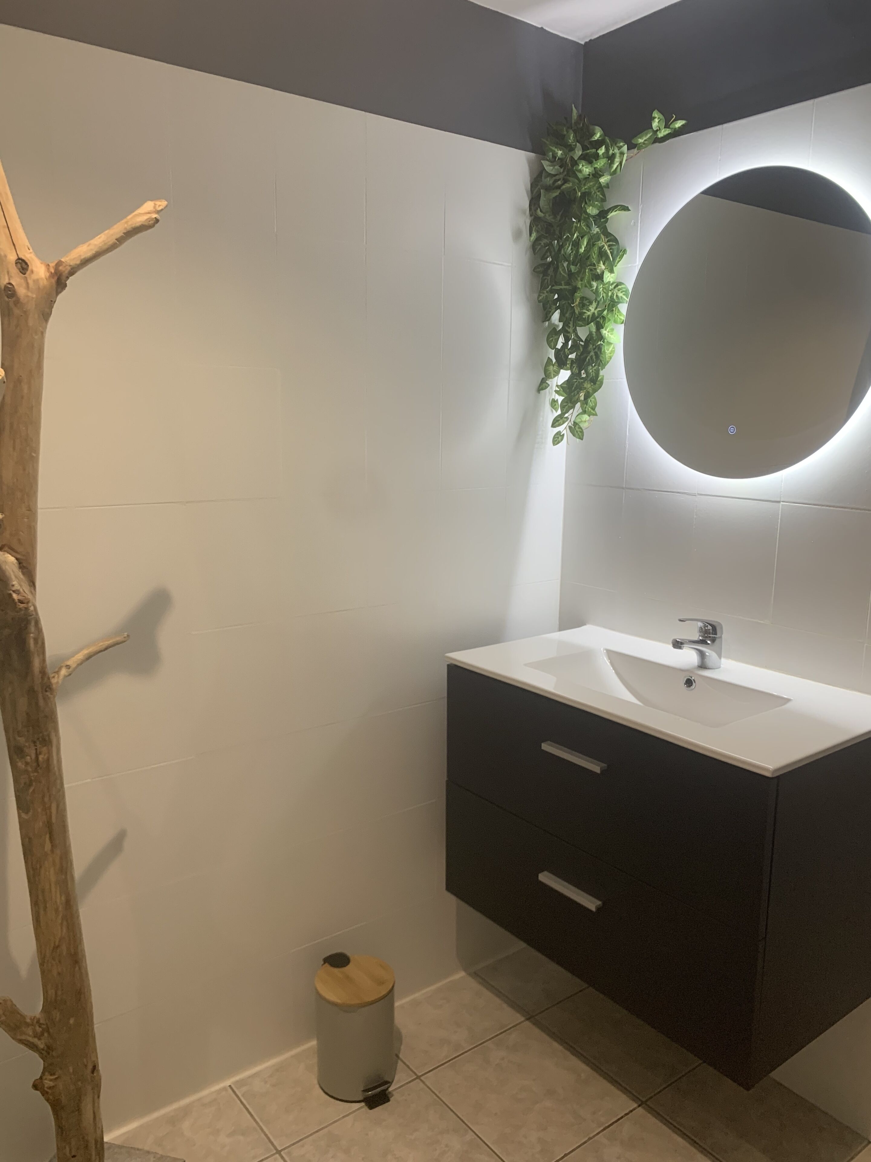 A modern bathroom with a white sink, dark cabinet, and a round mirror with a light.