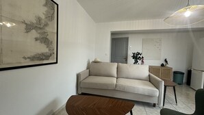 Una sala de estar con un sofá, una mesa de centro y una silla. Se observa una obra de arte enmarcada en la pared, una lámpara colgante y una puerta al fondo.
