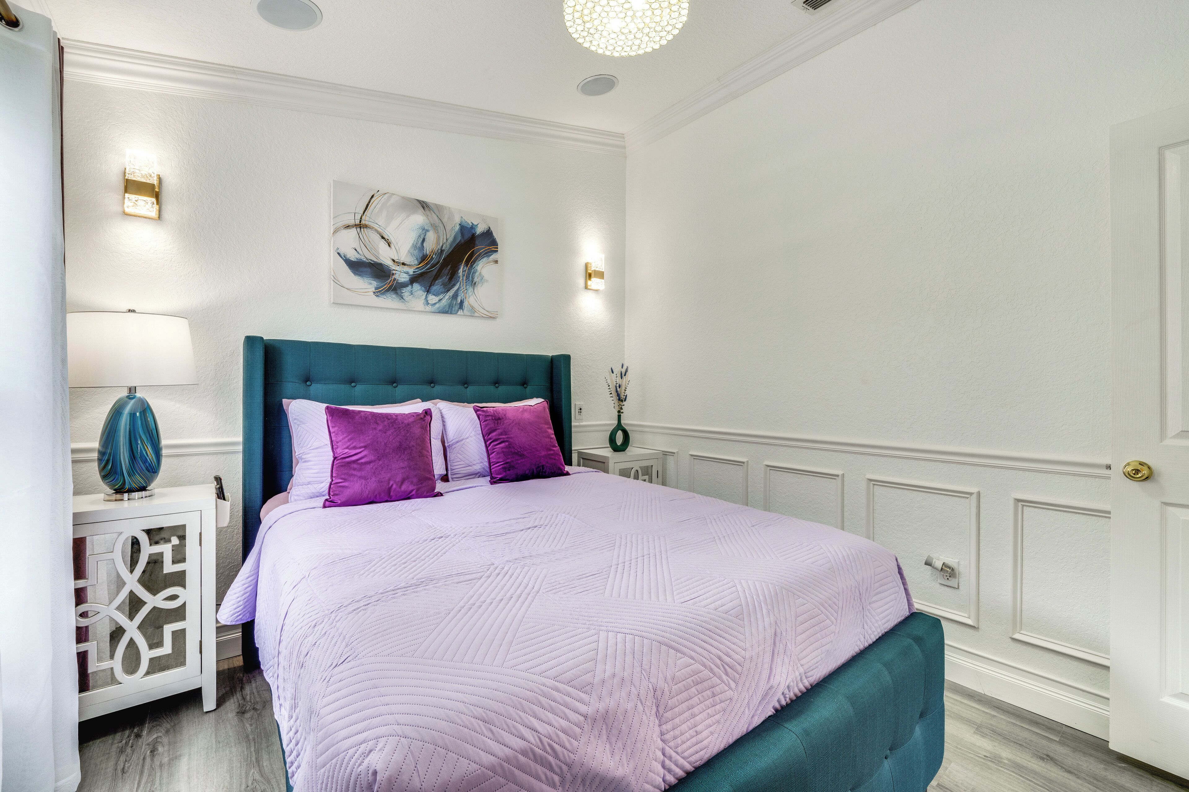 A bedroom with a teal headboard, a purple quilted bedspread, and a white wall with a painting.