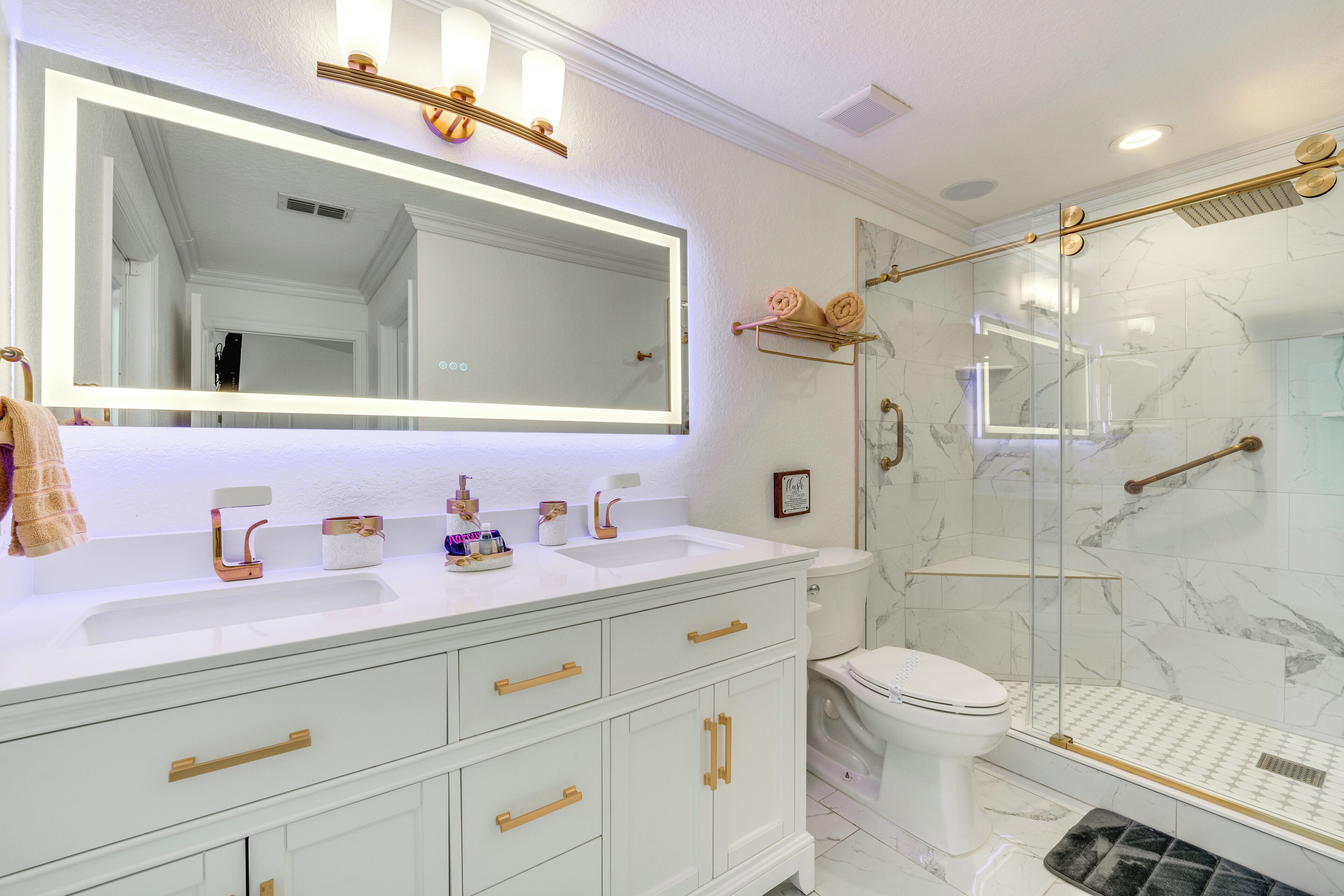 A modern bathroom with a large mirror, white vanity, gold handles, and a glass-enclosed shower.