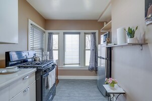 Cocina compacta con estufa, horno y refrigerador. Hay ventanas con cortinas, una mesita con un florero, y una repisa con objetos.