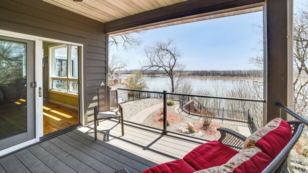 A covered outdoor deck with a view of a lake, a wooden ceiling fan, and a glass door leading to an interior space.