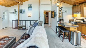 A cozy living room with a sofa, a wooden ceiling, a kitchen area with bar stools, and a staircase leading to another room. - Enchanted Woodland Cabin Near Yosemite w/ Fire Pit (Groveland)