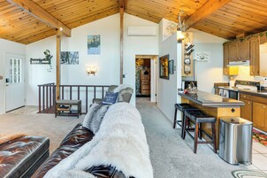 A cozy living room with a sofa, a wooden ceiling, a kitchen area with bar stools, and a staircase leading to another room.